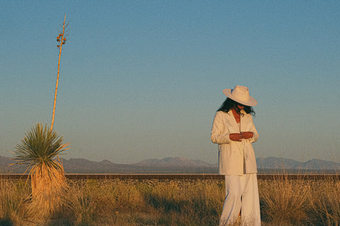 A woman in a white fringe blazer, white skirt, and white hat stands in the desert among dry grass and a dry agave plant. She is looking down and there are mountains in the distance.