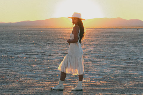 A woman with long dark hair stands in a dry desert playa in front of a sun setting behind mountains in the distance. She is wearing a white dress, boots, and hat, and is looking at the camera.