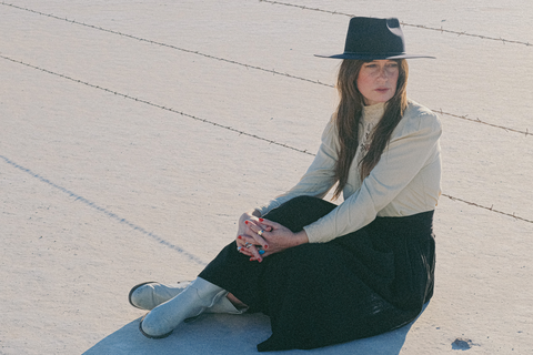 A woman with long brown hair sits on a dry playa in a cream and black dress, navy felt hat, and white boots. There is a barbed wire fence behind her.