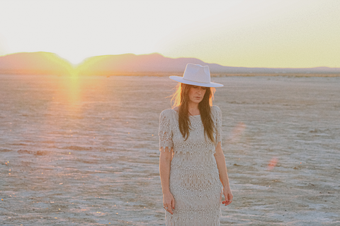 A woman stands facing the camera but looking down, wearing a white dress and white felt hat. Behind her is a dry desert playa and the sun setting behind a mountain.