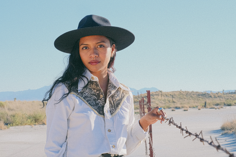 A woman with long curly brown hair stands touching a barbed wire fence in front of a dry desert playa. She is wearing a western brown and white button down shirt and a navy felt hat and is looking at the camera.