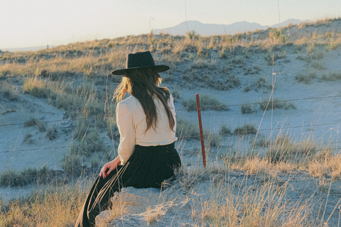 A woman sits on a hill in front of a dry rolling desert landscape. She is looking away from the camera and is wearing a cream and black dress with a navy felt hat.