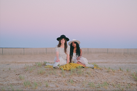 Two woman sit facing the camera on a chartreuse blanket on a small hill in front of a purple sky after the sun has set. They are both wearing white dresses and felt hats, one navy and one white.