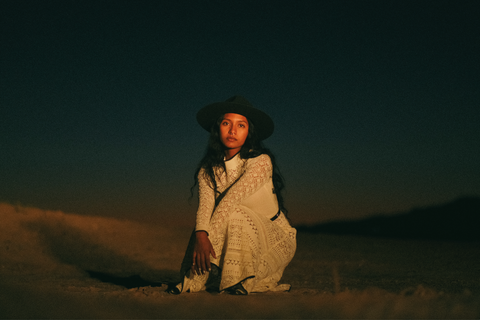 After dark, in yellow light, a woman crouches in a white crocheted dress and navy felt hat, looking at the camera.