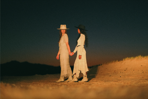 After dark, in yellow light, two women are holding hands, walking away as the one in front turns to look at the other. Both are wearing white dresses. The woman in front is wearing a white felt hat and the other is wearing a navy felt hat.