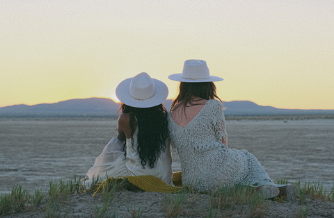 Two woman sit on a small hill facing a dry desert playa and mountains in the distance just after the sun has set. Both are wearing white dresses and white felt hats.