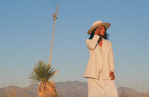 A woman wearing a white fringe blazer, white felt hat, and white skirt stands in dry desert next to an agave plant. There are mountains in the distance behind her and she is looking off to the right of the camera with her hand up by her face.