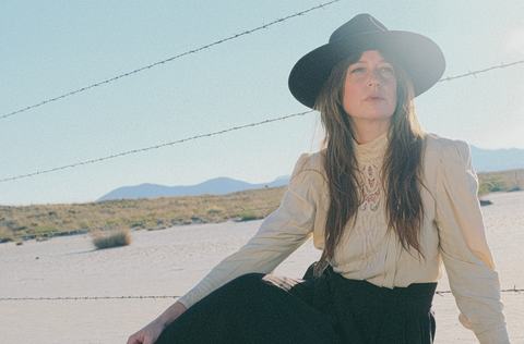 A woman with long brown hair sits on the dry desert plaza in front of a barbed wire fence. She is wearing a navy felt hat and cream and black dress and looking to the right of the camera.