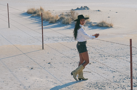 A woman stands facing a barbed wire fence with her head turned to the camera. She is standing in a dry desert playa and wearing a white and brown western shirt, black denim shorts, a navy felt hat, and tan cowboy boots.