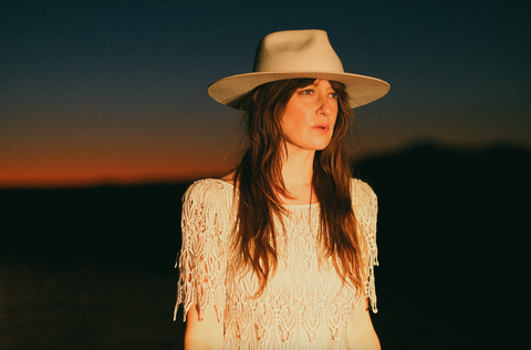 A woman with long brown hair stands in bright yellow light looking to the right of the camera. She is wearing a white dress and a white felt hat and the sun is set orange on the horizon behind her.