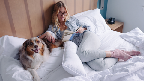 A woman reads while laying in bed with her dog
