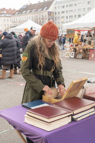 Beth wearing an orange knit hat and green army jacket digging through a box at a flea market in Vienna.