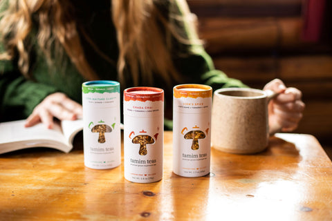 Assorted containers of Tamim Teas on a light colored wooden table in front of natural light in a well lit room.