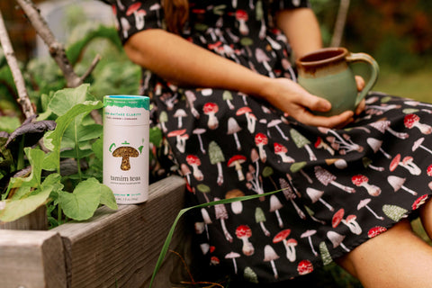 A container of Tamim Teas Lion-Maitake Clarity on a ledge next to a person in a dress sitting by a plant