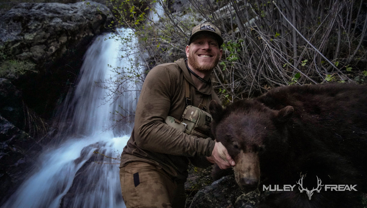 Erik Van Woerkom sitting next to a brown colored black bear that he shot in Idaho next to a beautiful waterfall