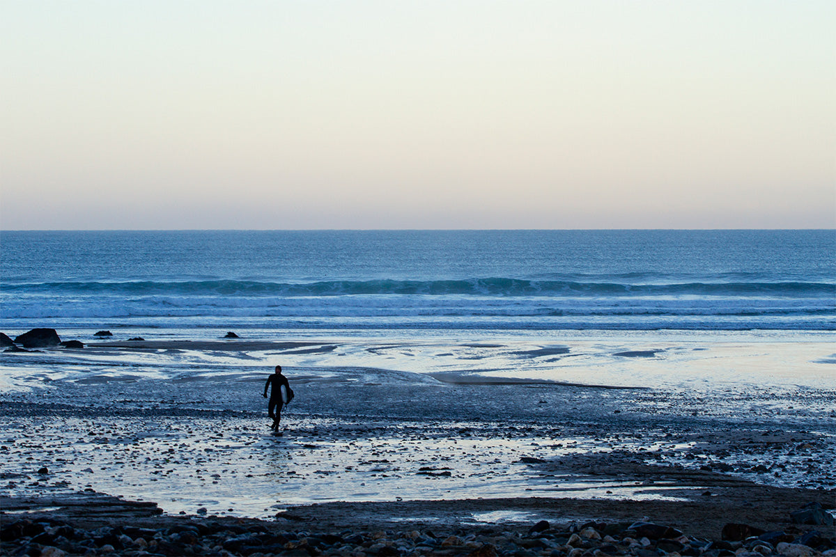 lone surfer walks back up the beach at dusk from the water