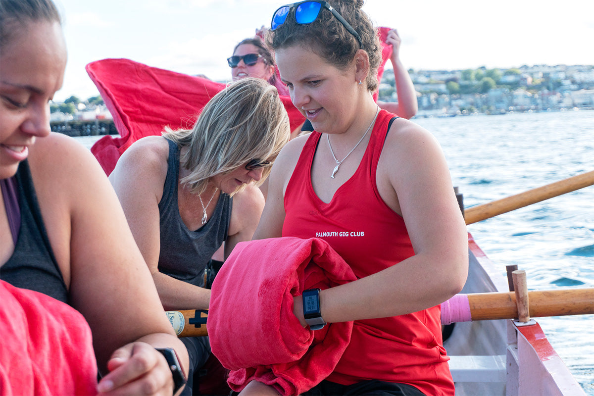 woman wearing red vest top reading Falmouth Gig Club rolls up her towelling robie 