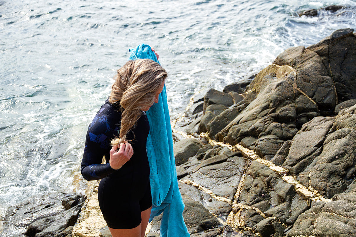 blonde woman takes her hair down wearing a short wetsuit holding a blue towel