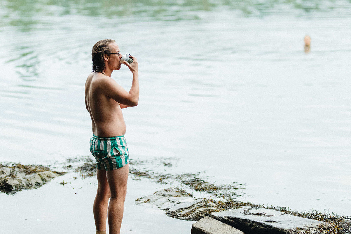 older man in green and white striped board-shorts sips a mug of coffee at the waters edge