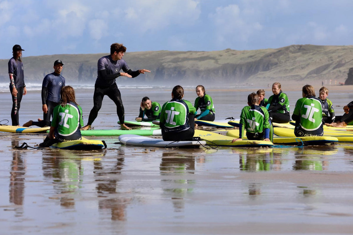 man in purple rash vest teaches class of young people to surf on the beach