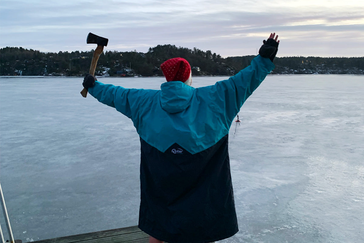 Sara stands by a frozen lake wearing a swim robe holding an axe