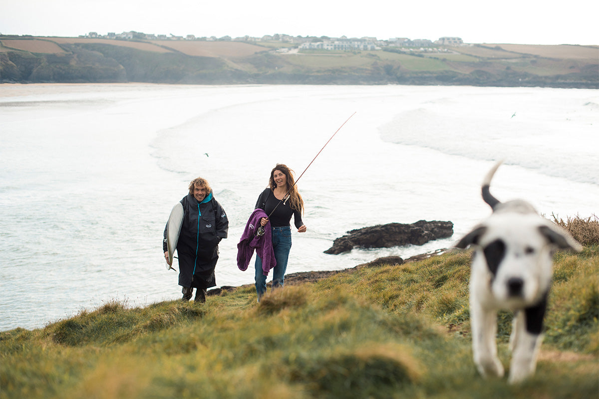 couple with fishing rod and surfboard follow black and white puppy away from ocean