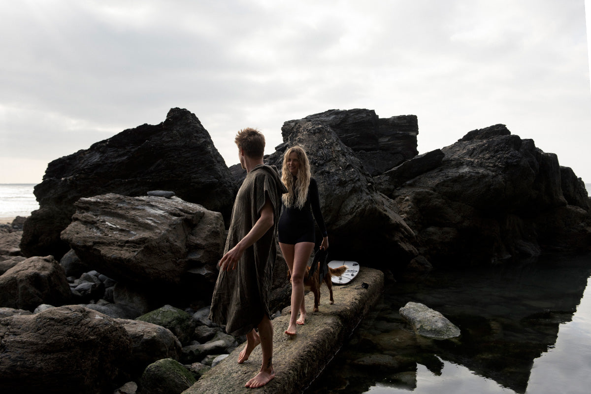 Blonde man and woman with spaniel walk along wall of tidal pool in towel robe