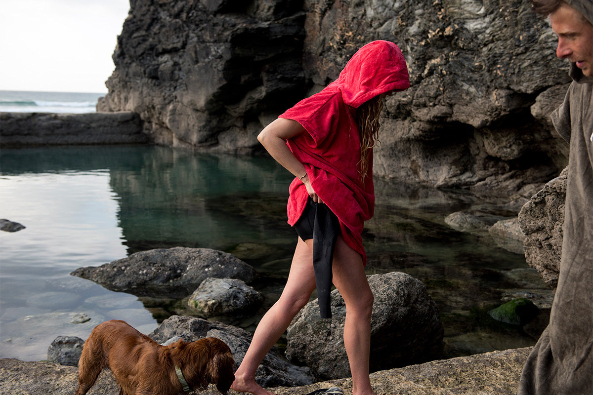 Blonde girl uses red towelling changing robe to get out of her short wetsuit by tidal pool edge