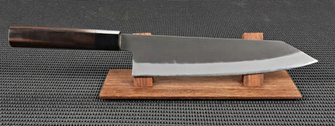 Hinoura Aogami Super Handmade 210mm Kiritsuke or K Tip Gyuto (chef/Cook) Japanese Kitchen Knife with a traditional Ebony handle and a Kurouchi (rustic/blacksmiths) finish on a red wood stand