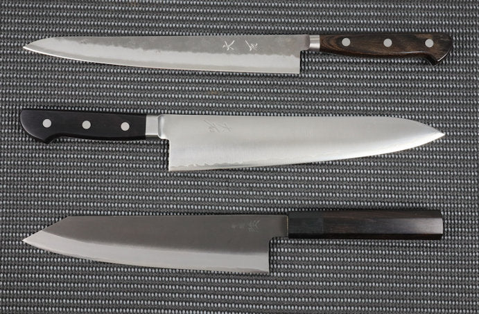 Three Hand made Japanese kitchen knives, all priced over $200, arranged vertically. The top knife is a Sujihiki (slicer) from the Ohishi Ginsan series. It has a long, 240mm, narrow blade with a fairly flat profile and a rough or pear skin finish. The middle knife is a Gyuto (Chef/Cook) kitchen knife with a polished 240mm blade that has a curved profile from heel to tip. The blade has an integrated bolster attached to a black Pakka wood handle with 3 rivets. The bottom knife is a Kiritsuke (Chef/Cook) kitchen knife. Its 210mm blade has a flatter profile from heel to its angular tip. The finish is the traditional dark Kurouchi or blacksmiths finish.