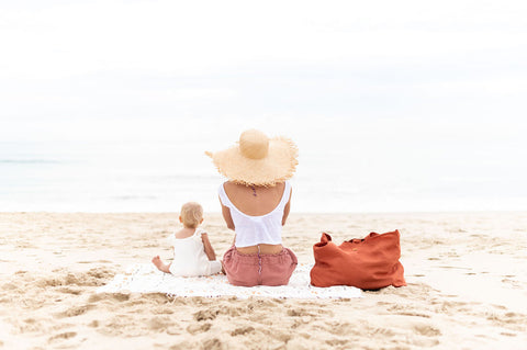 Mother and baby sitting on towel, over looking the ocean.  rust coloured pure linen bag placed beside the mother