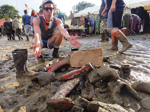 a man squatting next to a pile of lost muddy shoes at Glastonbury with a sign that reads, “Lost Souls”