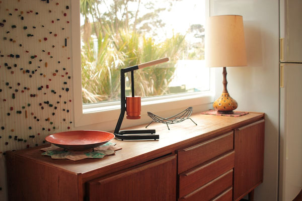 Lamp and coffee maker sitting on a mid-century sideboard by window.