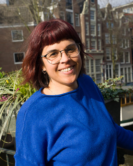Photo shows Ayelén looking into the camera and smiling. She is wearing an electric blue sweater. In the background are some Amsterdam's houses and some plants.