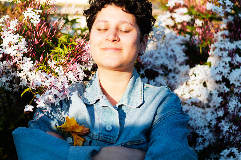 Victoria, the Stephen & Penelope staff photographer stands against a blooming bush with the sun in her face. Her eyes are closed and she is smiling