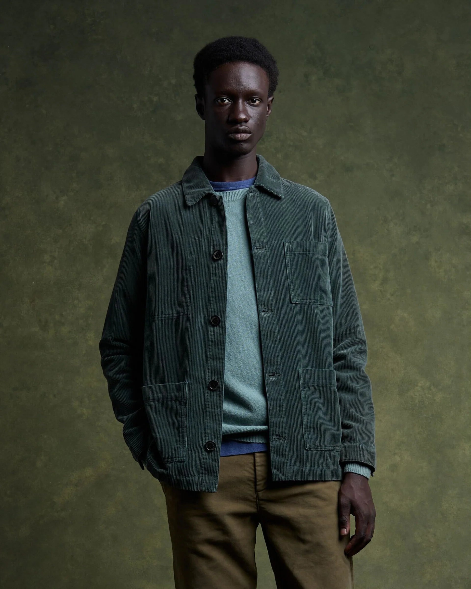 A person wearing a green corduroy jacket and olive pants against a green textured background.