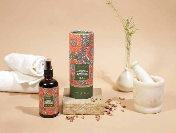 a beautiful photo of Manjish Glow Elixir - iYURA's bestselling night oil - accompanied by its beautifully illustrated, protective cylinder packaging