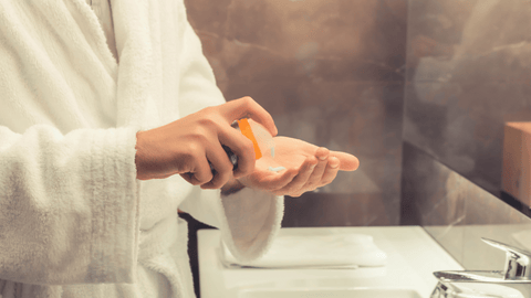 En närbild på en persons händer som trycker ut after shave från en orange flaska på handflatan. Personen bär en vit badrock och står framför en mörk marmorvägg i ett badrum.