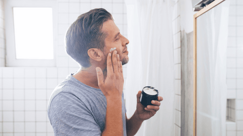 En man i en grå t-shirt applicerar raklödder på sitt ansikte medan han ser sig i spegeln, förmodligen förbereder han sig för att raka sig. Bakgrunden visar en ljus och luftig badrumsmiljö med vita kakelväggar.