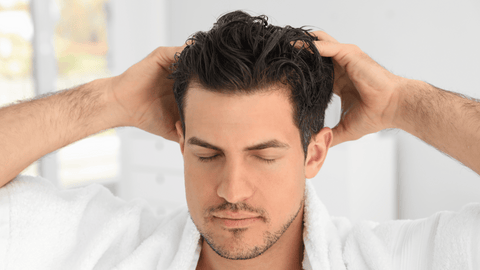 Bilden visar en man med vått hår som applicerar balsam eller hårinpackning. Mannen har stängda ögon och verkar vara avslappnad i processen. Han är insvept i en vit handduk, vilket antyder att han befinner sig i ett badrum.