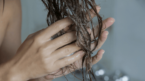 Bilden visar en närbild av en persons händer som applicerar balsam eller behandling i vått hår. Man kan se textur och glans i håret som tyder på vårdande produkter.