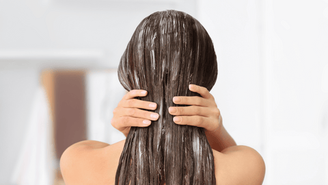 Bilden visar en person bakifrån som applicerar balsam eller hårinpackning i sitt långa, bruna hår. Personens hår ser vått och behandlat ut, vilket tyder på att de är mitt i en hårskötselrutin.