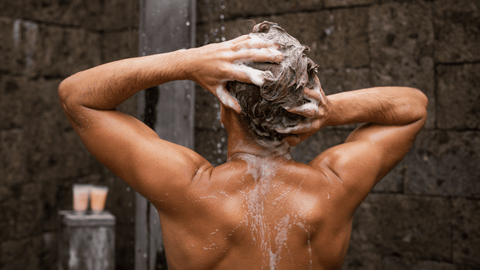 Bilden visar en person sett bakifrån som tvättar sitt hår och masserar in schampo. Personen är bar överkropp och står i en dusch med vattendroppar synliga i bakgrunden. Huden är våt och skimrar, vilket antyder att duschens vatten är på.