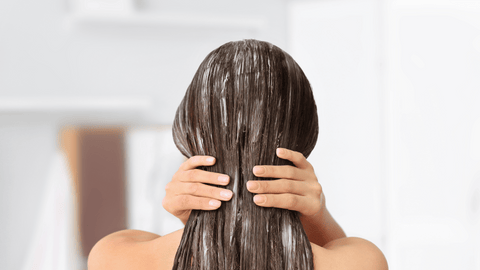 Bilden visar en person bakifrån som applicerar balsam i sitt långa, bruna hår. Håret ser vått och behandlat ut med balsam jämnt fördelat från rot till topp. Personens händer håller om håret vilket antyder att de arbetar in produkten genom håret.