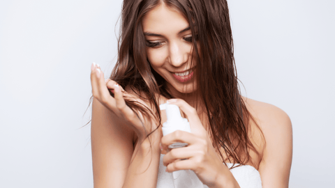 Bilden visar en leende kvinna som applicerar en hårvårdsprodukt på sitt hår från en pumpflaska. Hon bär en vit handduk, vilket tyder på att hon kan vara i en badrums- eller skönhetsvårdsmiljö.