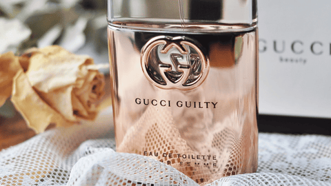 En flaska Gucci Guilty parfym på en vit yta med en suddig gul ros i bakgrunden och en vit textil under.