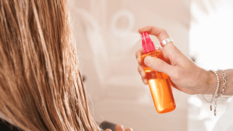 Bilden visar en närbild på någons hår där hårspray appliceras. En hand håller en orange sprayflaska med en rosa spraymunstycke. En fin dimma av sprayen syns i luften.