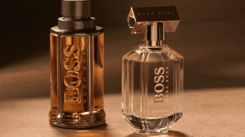Två eleganta BOSS-parfymflaskor, en klar och en gyllene, står på en mörk, texturerad yta.