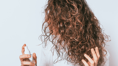 Person med böljande, lockigt hår håller en sprayflaska, redo att styla sitt hår, mot en vit bakgrund.