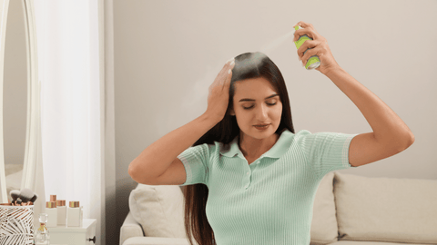 En kvinna med långt, rakt hår sprayar torrschampo på sitt huvud. Hon har en avslappnad hållning och en fokuserad blick, medan hon noggrant applicerar produkten.